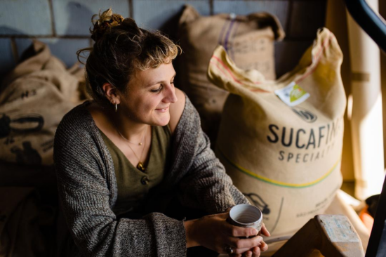 Noni sits in front of huge bags of green coffee from Sucafina and holds a ceramic coffee mug.