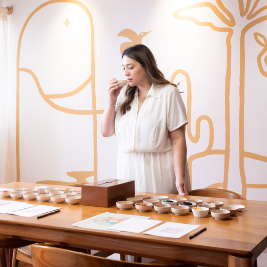 Maria holds up a small vessel to smell in front of her sniffing station set up on a sleek wooden table with matching chairs. She wears a white collared dress.