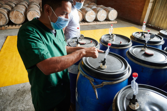 A coffee worker points to the nozzle on a blue tank roughly half his height. The room is full of wooden barrels stacked along the wall, and he stands next to a stand of blue barrels. The nozzle has a red cap to keep air out of the tank. Another worker observes. 