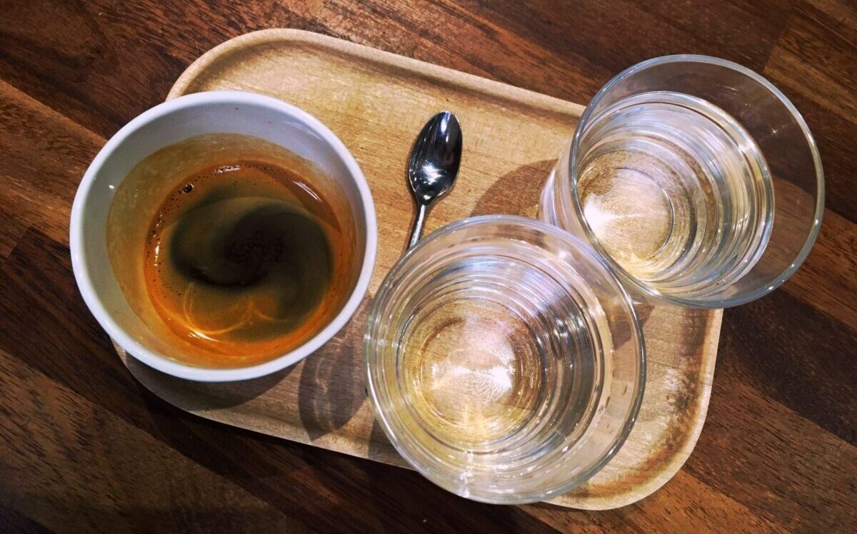 An espresso in white cup. There are are two small water glasses and an espresso spoon on the wooden tray.