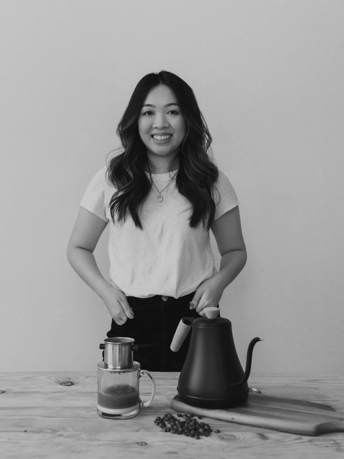Kim Dam in a black and white portrait with hands in jean pockets. She wears a necklace and white tee. In front of her on a large wooden table is a clear mug with Vietnames coffee brewing into it from a phin filter. There is also a pour over kettle.
