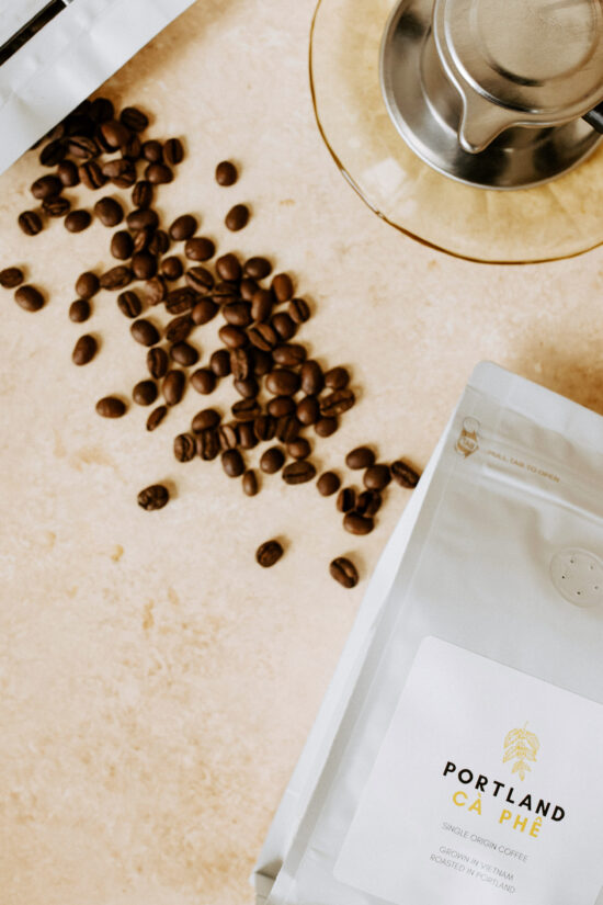 A bag of Portland Ca Phe single origin beans grown in Vietnam. The bag is white with a pull tab on the top. It has the company name in black and yellow caps on a sticker. On the surface nearby are spilled coffee beans and a metal phin filter.