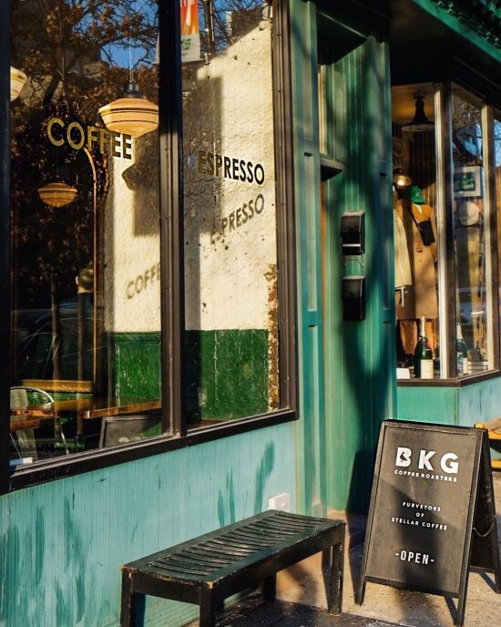 The front of BKG Roasters in Clinton Hill. There is a large window with Coffee and Espresso painted on the outside, and light fixtures and tables just visible inside. Outside the window is a black slatted bench and a black sandwich board sign that has the company logo, a B with a pigeon outline inside it and capital K and G.