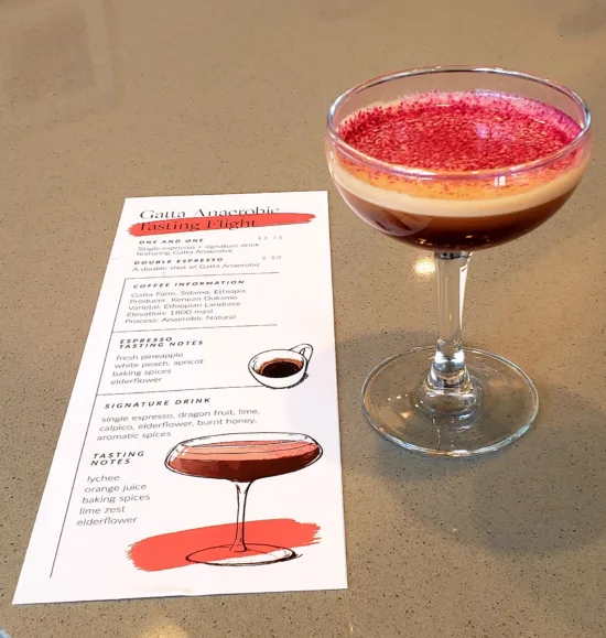 The Gatta Anaerobic drink flight, shown here with a martini glass and an info sheet. The signature drink is layered with espresso, milk, and pink topping.