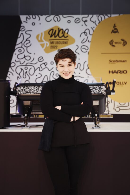Morgan Eckroth poses, arms crossed and smiling, in front of an espresso machine at the World Coffee Championships. They wear a black turtleneck and apron and have short black hair.