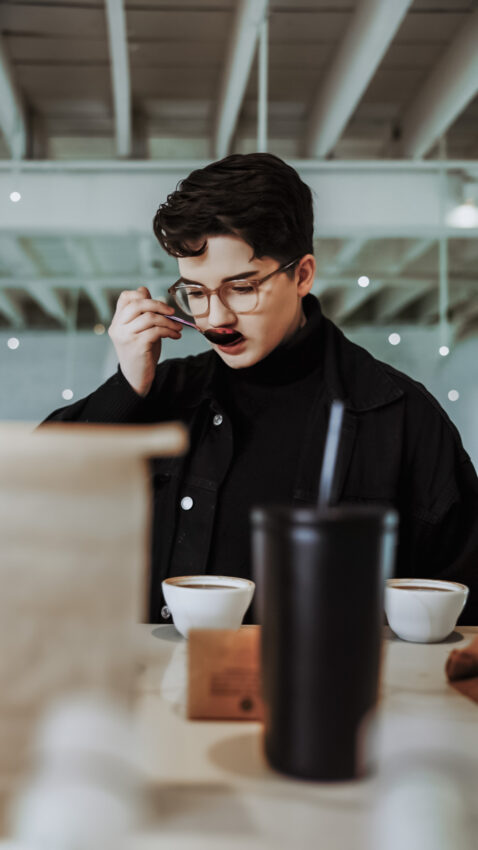 Morgan Eckrtoh sips from a rainbow spoon out of a white cupping vessel. They wear glasses and a black shirt. A black cold drink tumbler and kraft paper coffee bag are faded in the foreground.