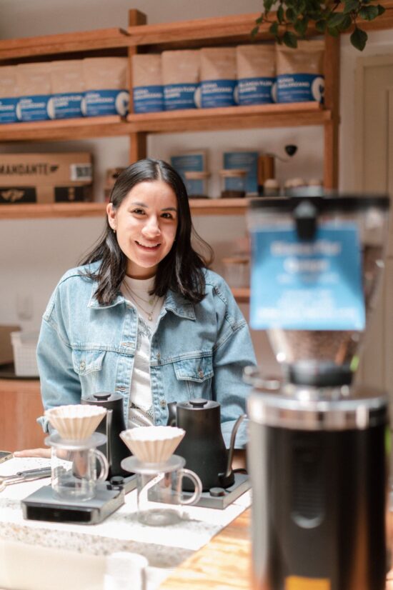 Carolina stand behind the espresso bar about to brew pourovers with Fellow kettles. She wears her hair down and has on a light blue denim jacket.