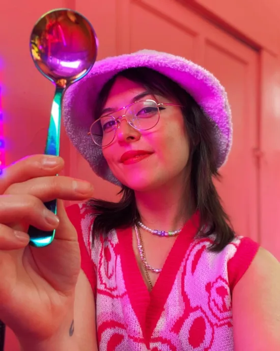 Umeko holds up a metallic rainbow cupping spoon. They wear wire rim glasses, a fuzzy bucket hat, and beaded necklaces over a sweater vest. The lighting is pink.