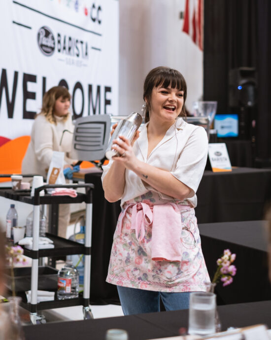 A barista competitor wears a pink floral apron and shakes a cocktail shaker with ingredients for her beverage. The coffee set up includes matching pink flowers, info cards and clear water glasses.