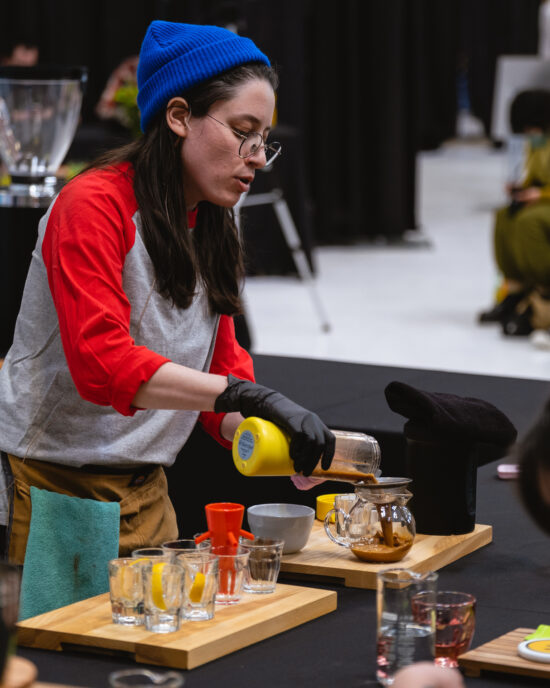 A barista in bright colors pours a coffee mixture from a blender into a glass server, while discusiing the story behind the drink.