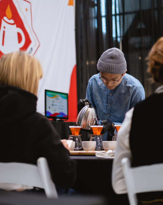 A barista in the brewer's cup category pours water from a silver kettle into a pour over. He wears a gray beanie, glasses and denim shirt.