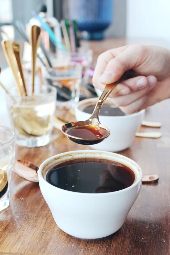 A hand with a cupping spoon has dipped coffee out of a ceramic cupping vessel. In the background, there are spoons sitting in water glasses for rinsing.