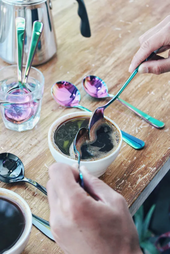 Two hands hold rainbow cupping spoons over coffee in a ceramic cupping vessel, breaking up grounds on top of the brewed coffee. On the table is also a small glass cup of water with two more spoons and a water kettle.