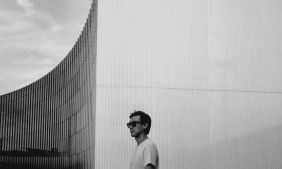 Black and white photo. Eric Park wears sunglasses and stands near the side of a long, curved glass building.