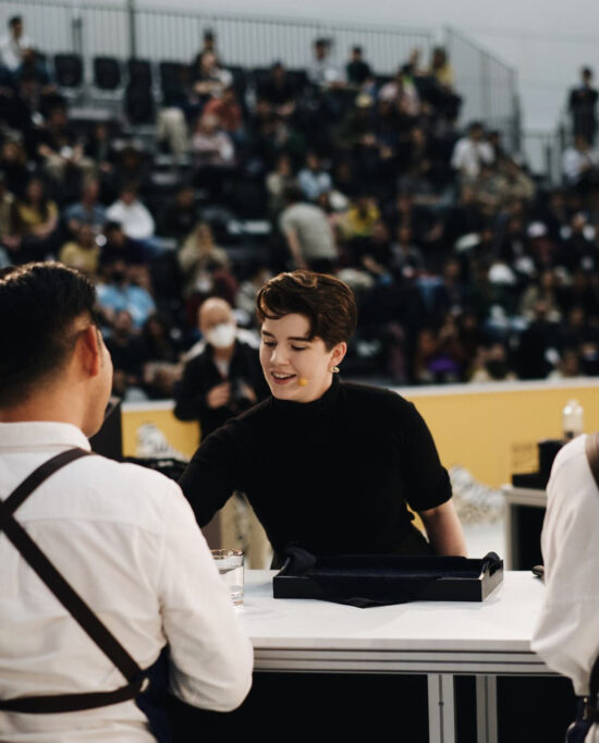 Morgan performs their routine in front of a large crowd in the background. They are in front of a competition table pouring for judges, with a black tray on the table.