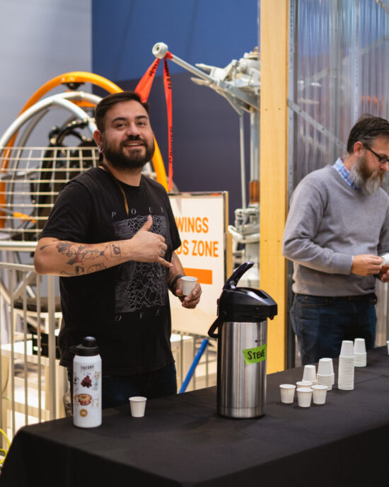A roaster in a black tee shirt gives a hang ten sign while offering coffee samples. The coffee is in a metal urn, and served in small paper cups. Museum paraphernalia is just visible behind the coffee table.