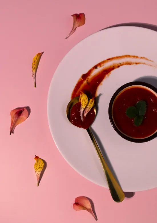 A gold spoon with sauce in the bowl and a small bowl with more sauce inside it and a sprig of mint, all on a round white ceramic plate. The background is pink, with colorful flower petals placed throughout.