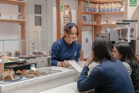 A barista in a blue sweat shirt and headband shows the menu to two seatede customers at the coffee bar. Beside her is a glass pastry case, and behind a shelf with clear glass mugs. In the corner, there are wooden shelves stocked with coffee and shop merch.