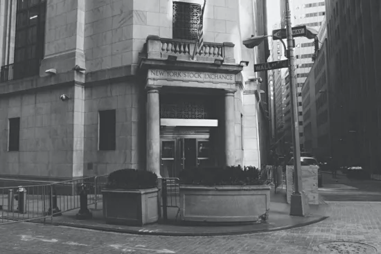 The marble/stone exterior facade of NY stock exchange, in black and white. It has a corner-facing entrance onto Wall Street, with cobbled stone streets in front. Three glass doors lead inside, one of which is open Two stone columns flank the sides of the entrance and NEW YORK STOCK EXCHANGE is chiseled into the stone sign above the doors. There is an American flag flying above the sign. Large planters with greenery are situated on the sidewalk in front.