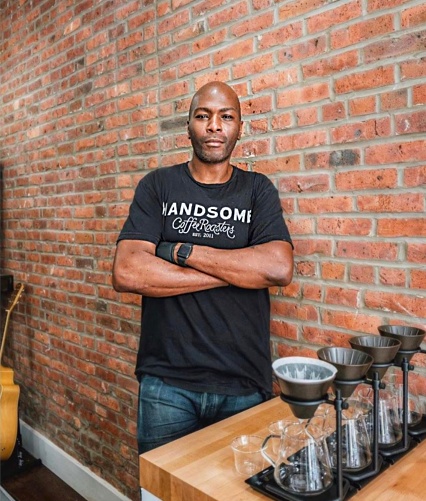 Nigel stands with arms crossed in front of an exposed brick wall, wearing a shirt that says "handsome coffee roaster." Four pour over setups sit on the wooden blovk counter in front of him, and there is an acoustic guitar leaning against the wall in the background.