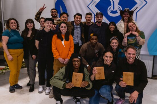 A huge group of 18 competitors, many in bright colors, one cowgirl hat, and three wooden plaques held up by winners crouching in front.