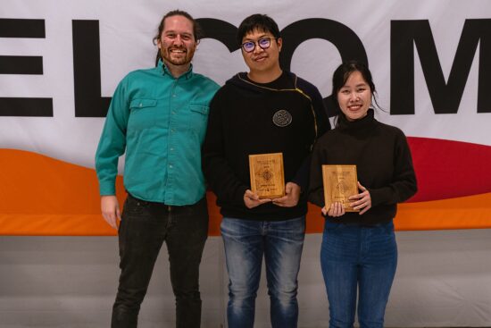 Piyapat is in a blue button up shirt, wenbo wears a side-zip hoodie, and Truc wears and brown turtleneck. They all wear jeans, two are holding up plaques.