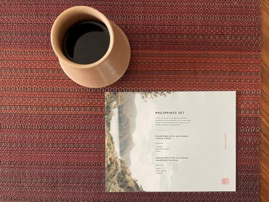 A red woven placemat with a ceramic coffee mug and a white printed paper describing the coffees (text too small to read). On the left side of the paper, sideways, is a lake with vegetated mountains behind it.