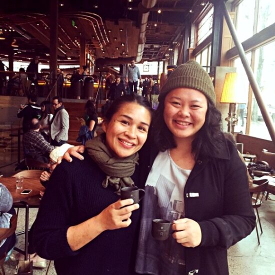 Two women pose with small black coffee cups. Blue Bottle's Selina Viguera on left wears a brown scarf and sweater, while Carmel on right has her arm around Selina's shoulder and wears a black jacket and brown knitted hat.