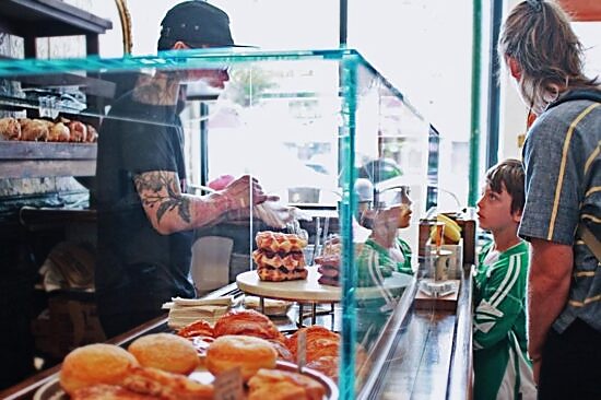 A tattooed barista serves two customers, an adult and child, at the pastry case inside BKG, which has donuts, waffles and croissants on serving plates.