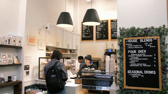 The bar area inside CPNY has black hanging lamps, a black Synessa espresso machine. and black felt menu boards. Retail shelving with coffee bags and brewing methods is to the left.