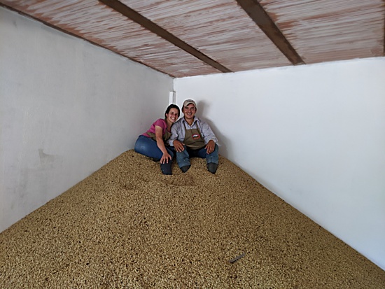 Helena and her husabnd sit in a corner atop an enormous pile of unroasted green coffee beans, both wearing brown aprons and smiling.