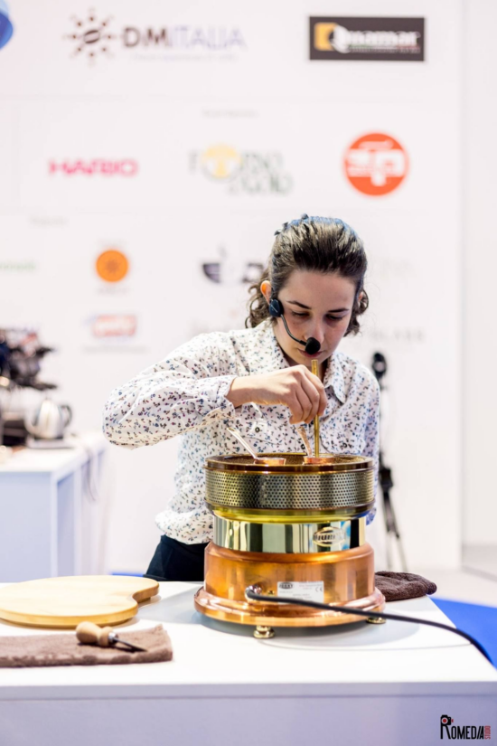 Helena wear a patterned shirt and stirs coffee in an ibrik at the Cezve/Ibrik Championship in Italy in 2016. She wears a headset. Blurred sponsor brand logos are visible behind her on a white wall behind the stage.