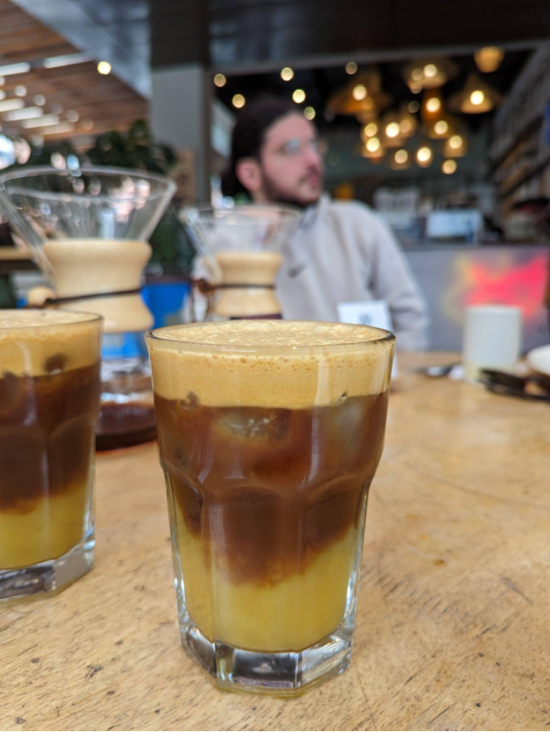 A layered coffee beverage in glass sits on a table at Azahar. The three layers: foamy on top, darker brown middle, lighter brown/tan on bottom. Blurred in the background are a person sitting on the other side of the table and the string lights on the shop ceiling.