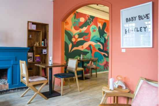 Inside El Pergolero, the walls are pink, the fireplace mantel is bright blue, and there is a coloful mural on the wall under the arch to the next room. Chairs are vintage mismatched wood, and there is a recessed modern bookcase in the wall with coffee supplies. A white markerboard sign reads BABY BLUE BENTLEY.