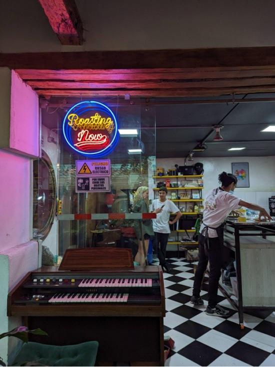 The inside of Jaguar has retro black and white checkered floors, a dual-keyboard  electric piano, yellow merch shelves and a colorful neon sign that reads Roasting Now.