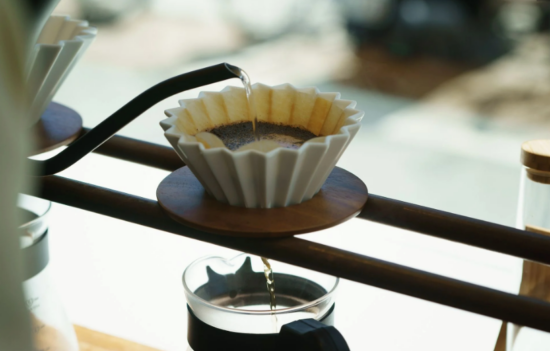 The Origami dripper is resting on top of two rounded bars on a holding device set above several decanters. The dripper is white ceramic with sharp textured ridges, similar to what you'd feel on paper filters for drip coffee.
The Origami has a wooden base attached to help stabilize it on the frame.