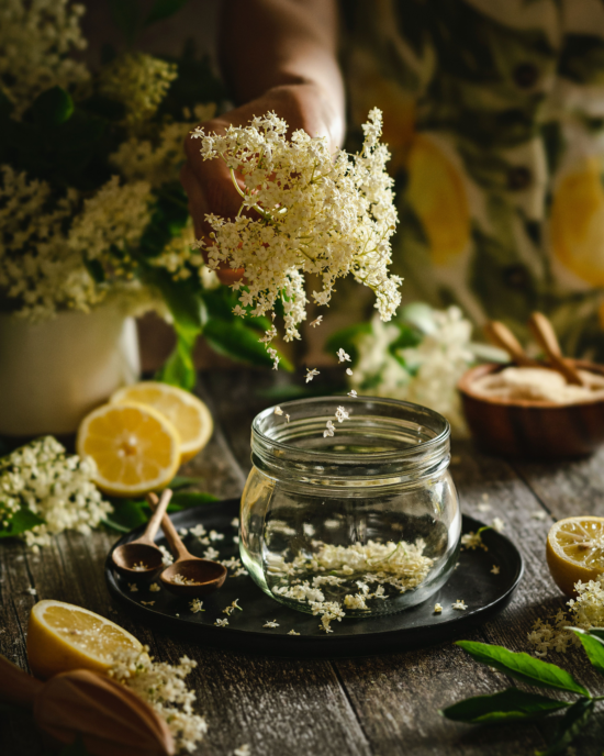 A wood table with a black plate. On the plate is a glass jar and elder flowers being placed into it, and two wooden spoons. Elderberry branches with flowers and leaves and lemons sliced in half adorn the table. 