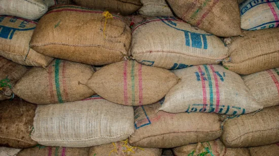 A big stack of rough fabric green coffee bags are piled up. They are mostly linen colored but have some colored stripes or labels on them.