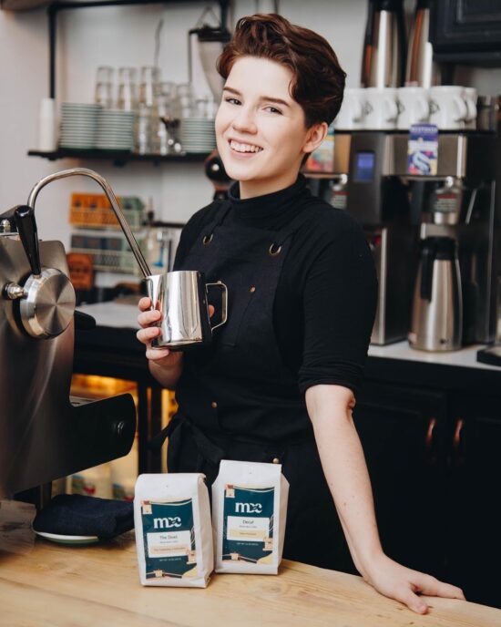 Morgan, steaming pitcher in hand and wearing a dark apron and black turtleneck, smiles at the camera behind an espresso machine. In front of them on the wooden counter is bagged coffee branded with Morgan Drinks Coffee logo.