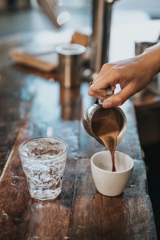 A woman's hand pours espresso from a metal pitcher into a white porcelain espresso cup, next to a small rocks glass of sparkling water. 