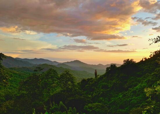 A sunset in Colombia over tree-packed, deep green mountains and small valleys. The sky is partly clouded and has notes of pink, orange and yellow fading up to blue.