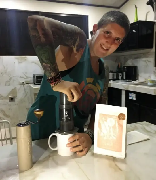A person with a shaved head and tattoos smiles while pressing an AeroPress into a mug in a hotel room. The X25 and coffee bag are also on the counter.