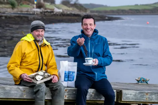 Alex and David drink coffee on a dock by the sea with a box of oysters and wear blue and yellow rain jackets.