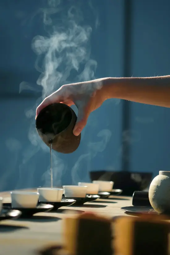 A hand pours brewed tea from a black pitcher into small white cups on black saucers. Steam rises up from the tea.