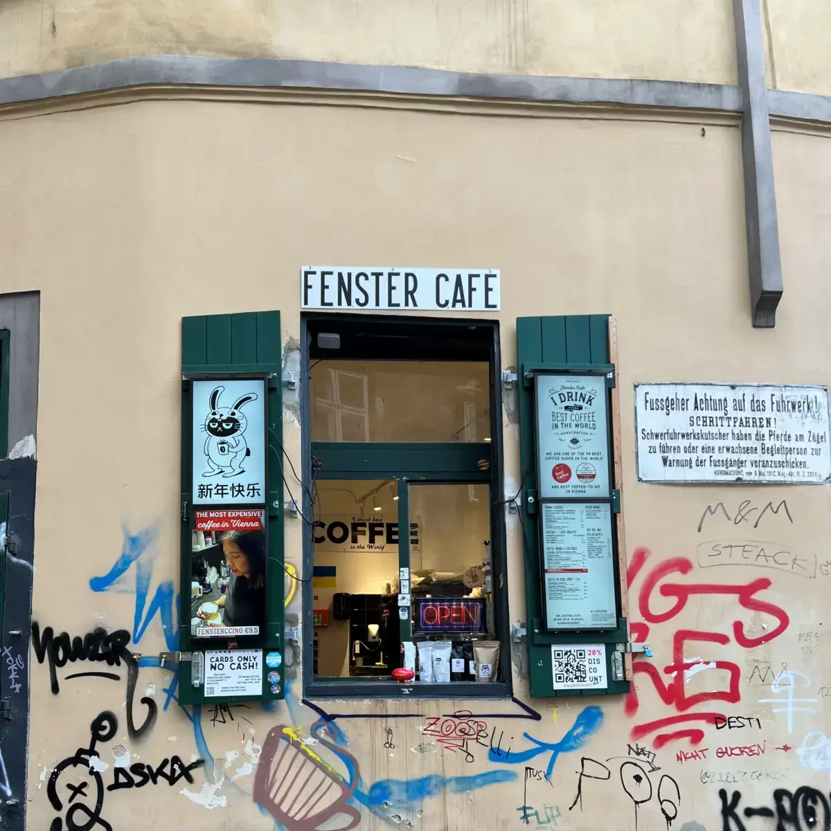 The window at Fenster. It has wooden shutters, and a sliding window. The menu is attached to the shutter. Graffiti tags cover the surrounding walls.