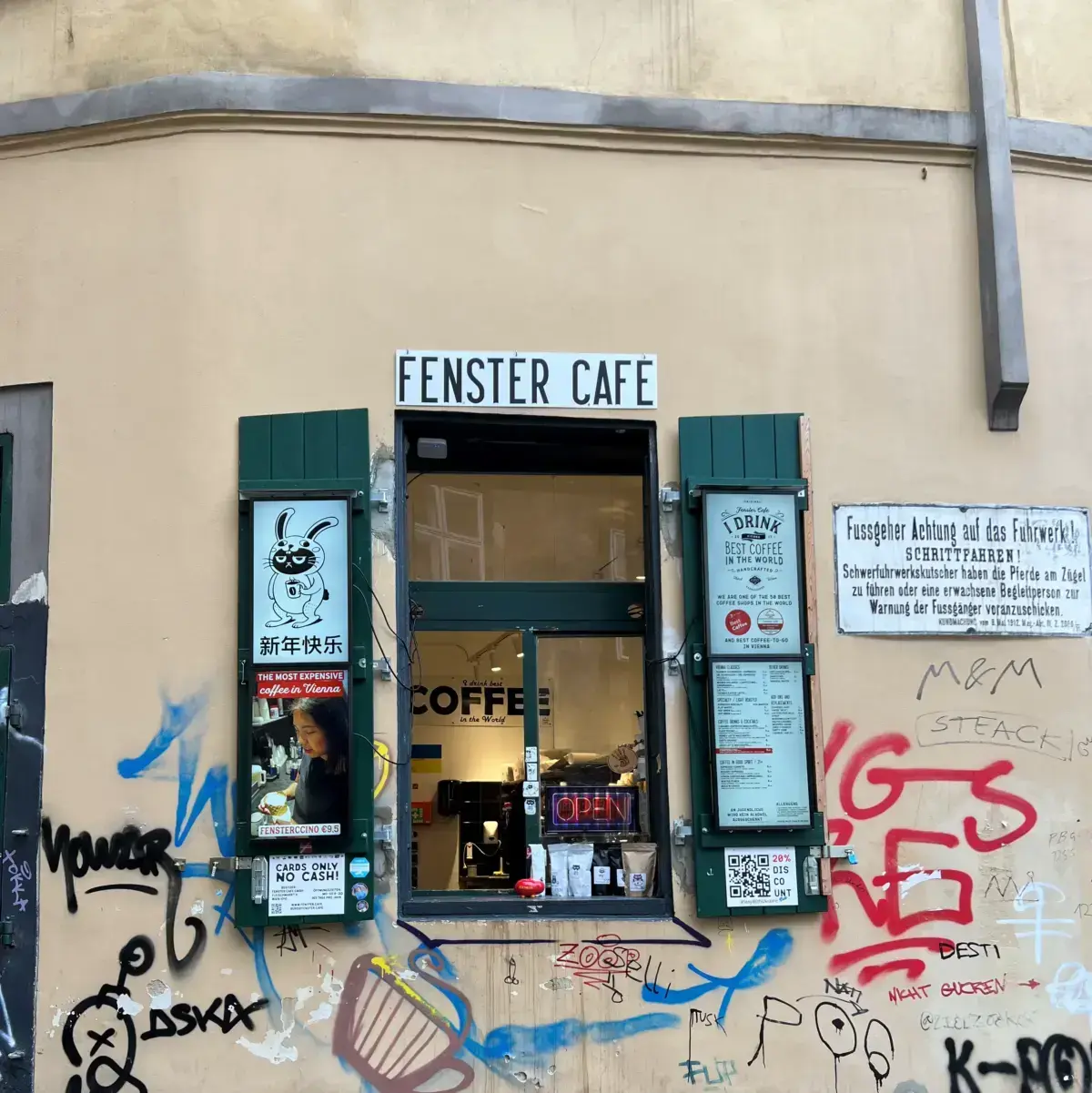 The window at Fenster. It has wooden shutters, and a sliding window. The menu is attached to the shutter. Graffiti tags cover the surrounding walls.
