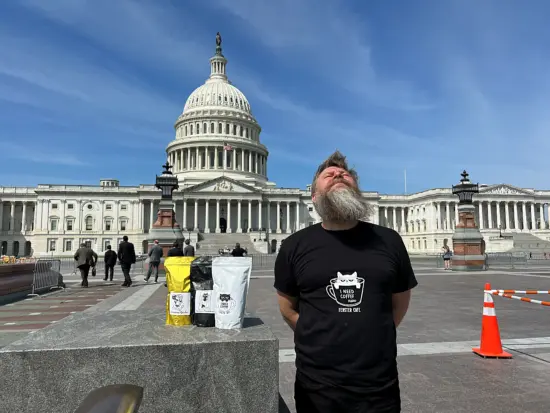 Sashko gets his photo taken in Washington DC with some coffee bags. He wears a t shirt with a cat in a coffee mug and stands in front of the US Capitol bulding, which is white with many windows and pillars and a huge done with statue on top.