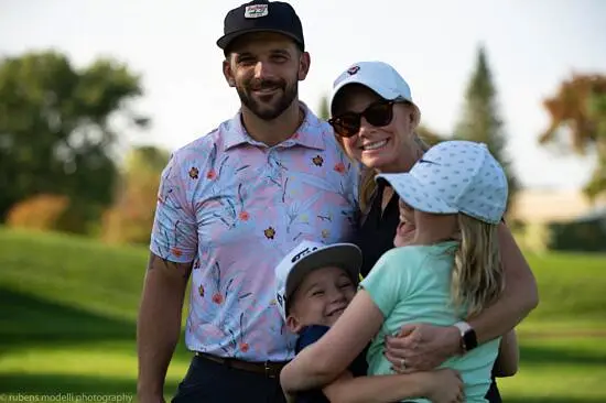 Noah with with his family: wife and two kids. The are all hugging and wearing ball caps and bright colors. They're standing outside on the grass, with trees in the background 