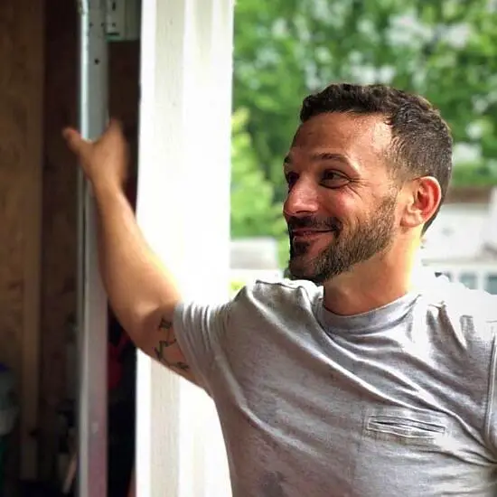 Noah Namowicz uses his experience with sobriety to help others stay sober. Here he stands in a doorway smiling, with one arm on the door frame, wearing a pocket t shirt.