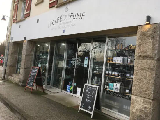 Le Café Qui Fume has a full glass front with sandwich board signs and the name painted on the white wall above. Retail coffee sits on the shelves inside. Residential windows are located above the shop.
